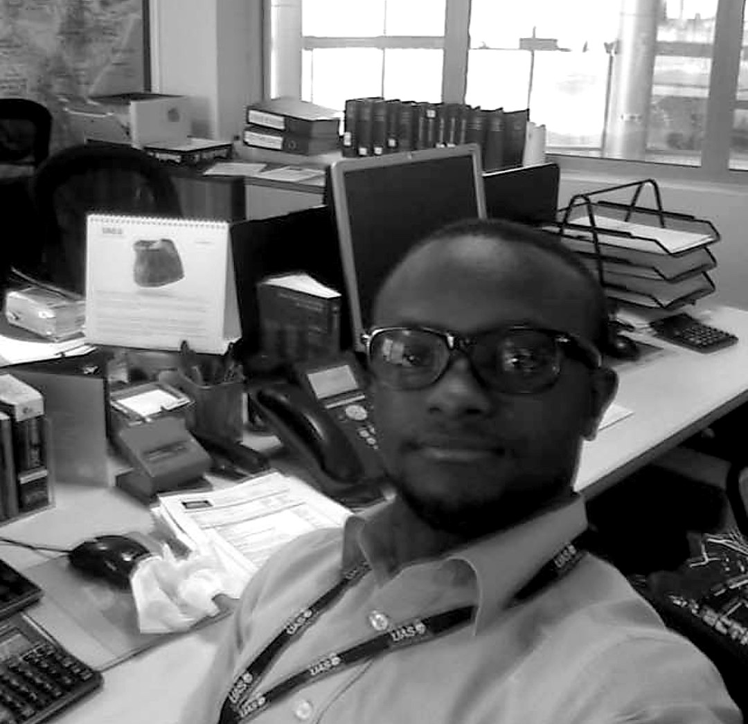 A person seated at an office desk with a computer monitor, stacked paperwork, a desk phone and a calculator, in front of a window.